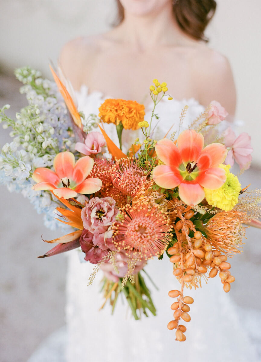 At her art-inspired wedding, a bride carried an asymmetrical bouquet of flowers in shades of peach, orange, pale blue, yellow, and pink. The blooms included reflexed tulips, pincushion protea, carnations, and birds of paradise.