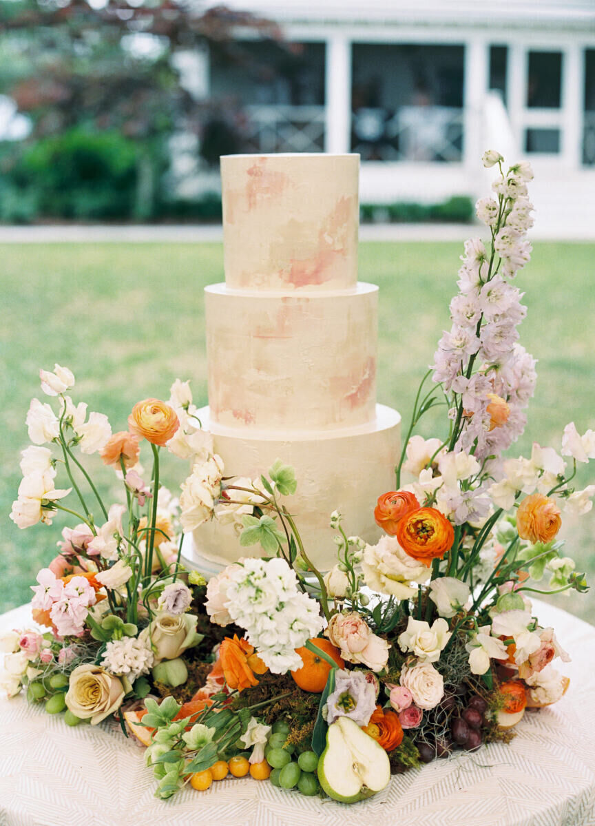 A three-tier wedding cake with a soft pink textured finish is surrounded by bright flowers at an elegant spring wedding.